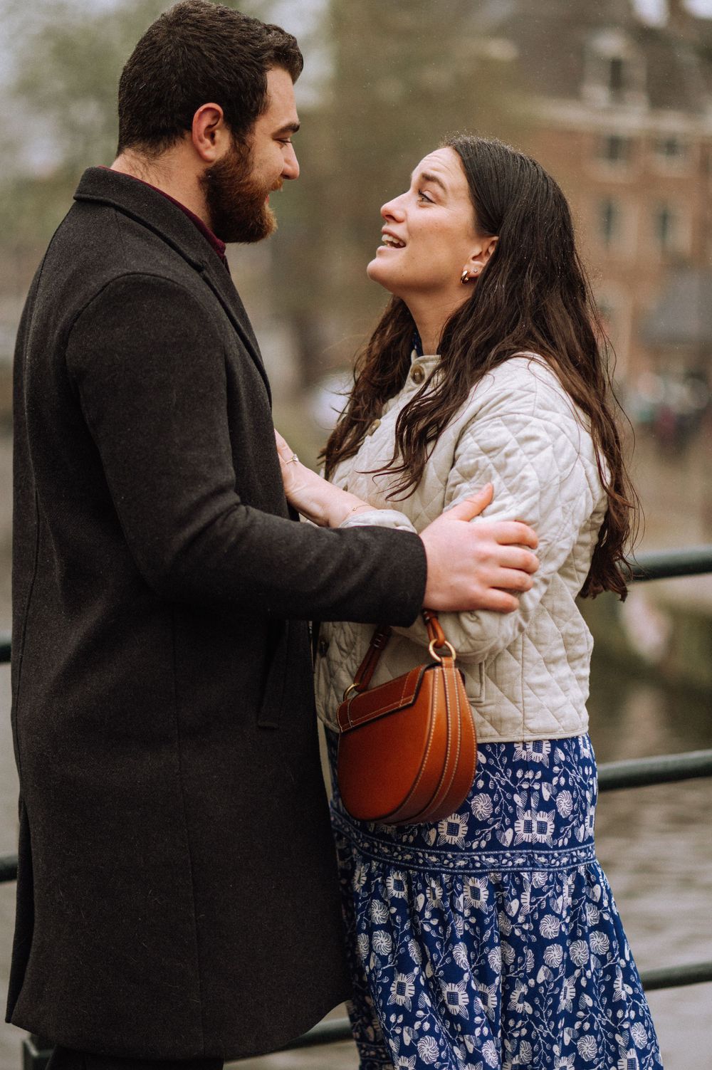 A couple embraces on a bridge in Amsterdam, wearing a black coat and floral dress.