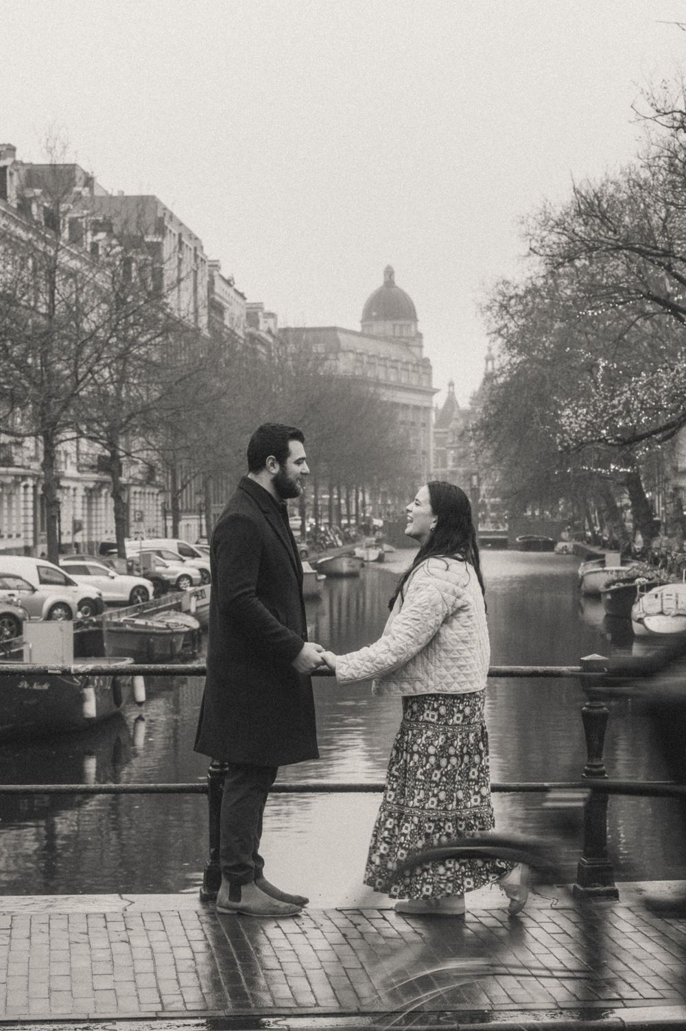 A couple stands along an Amsterdam canal with historic architecture in the background.