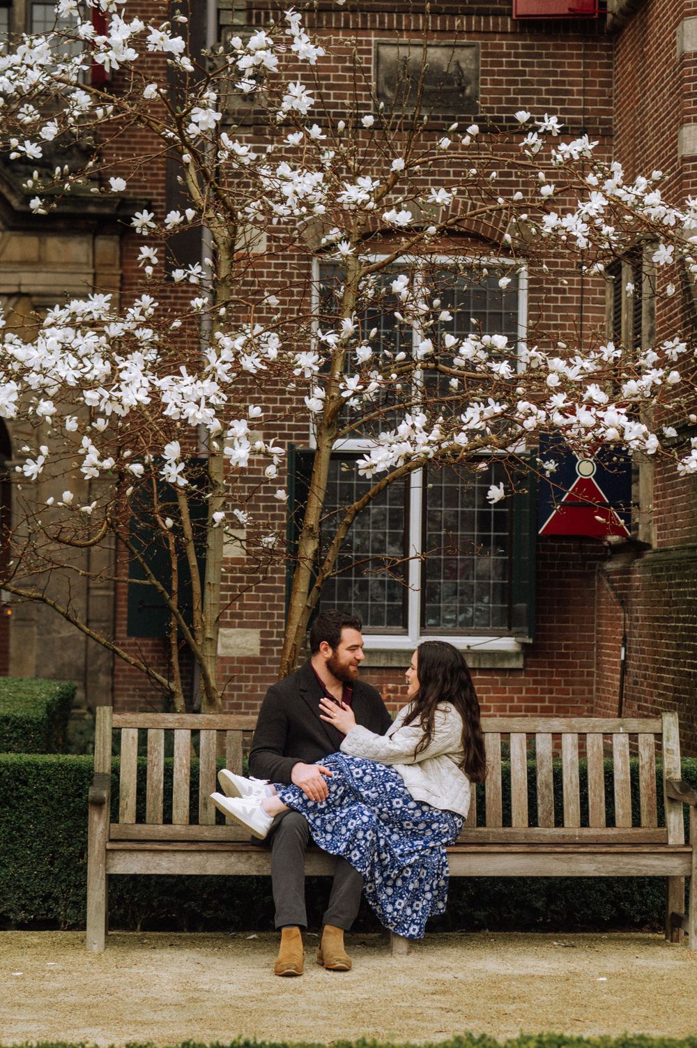 A couple sits on a wooden bench under blooming magnolia trees near a brick building.