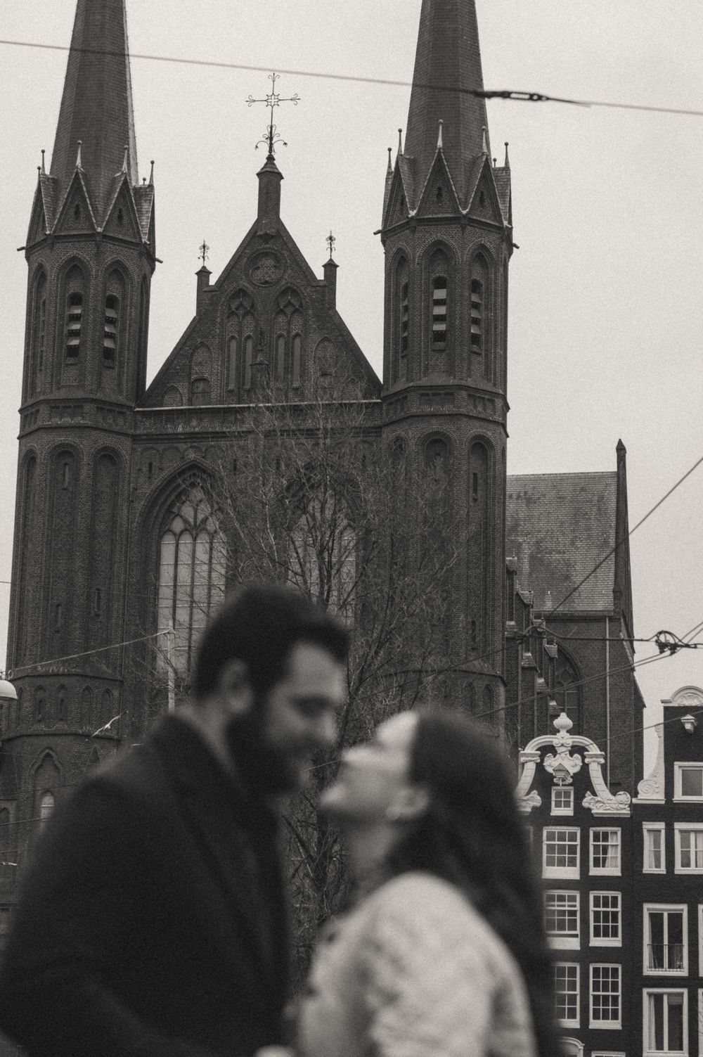 Blurred couple about to kiss in black and white in front of a Gothic twin-spired church and canal houses in Amsterdam.