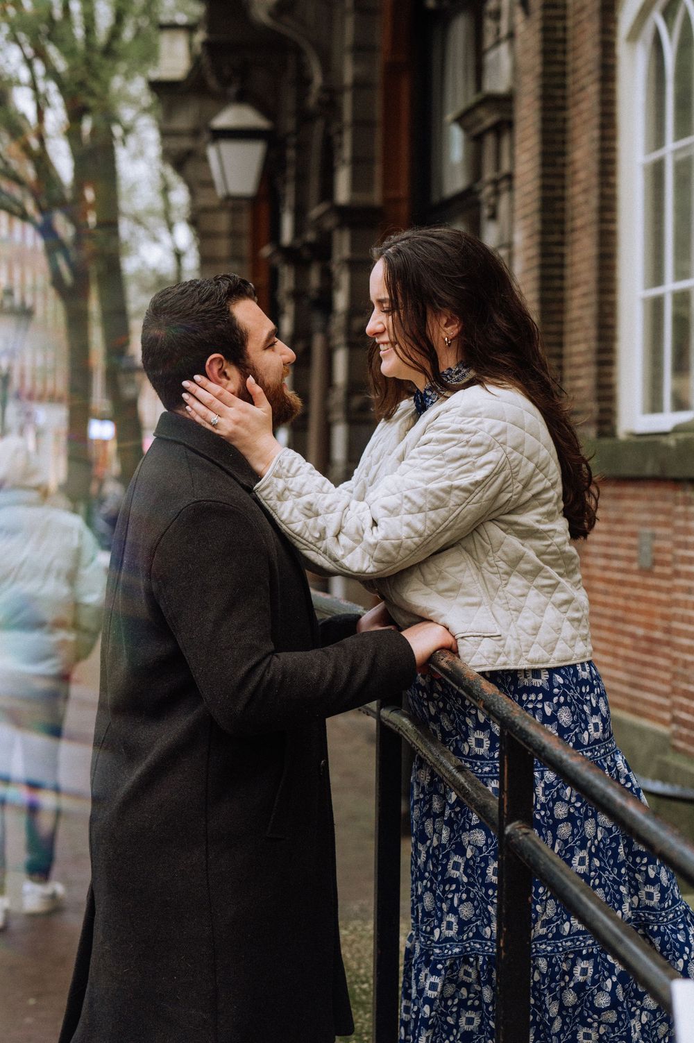 Smiling couple embracing beside a metal railing on a city street, the woman showing her engagement ring beside a brick building.
