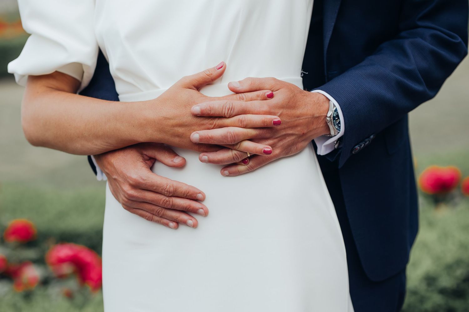 Close-up of bride and groom embracing, hands wrapped around her white dress near a rose garden.