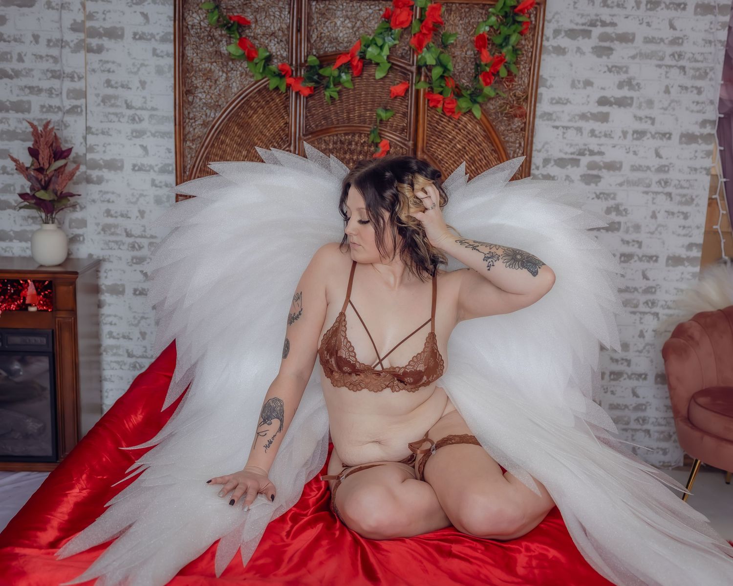 Woman in brown lingerie poses with white angel wings against brick wall backdrop with red roses and festive decor.