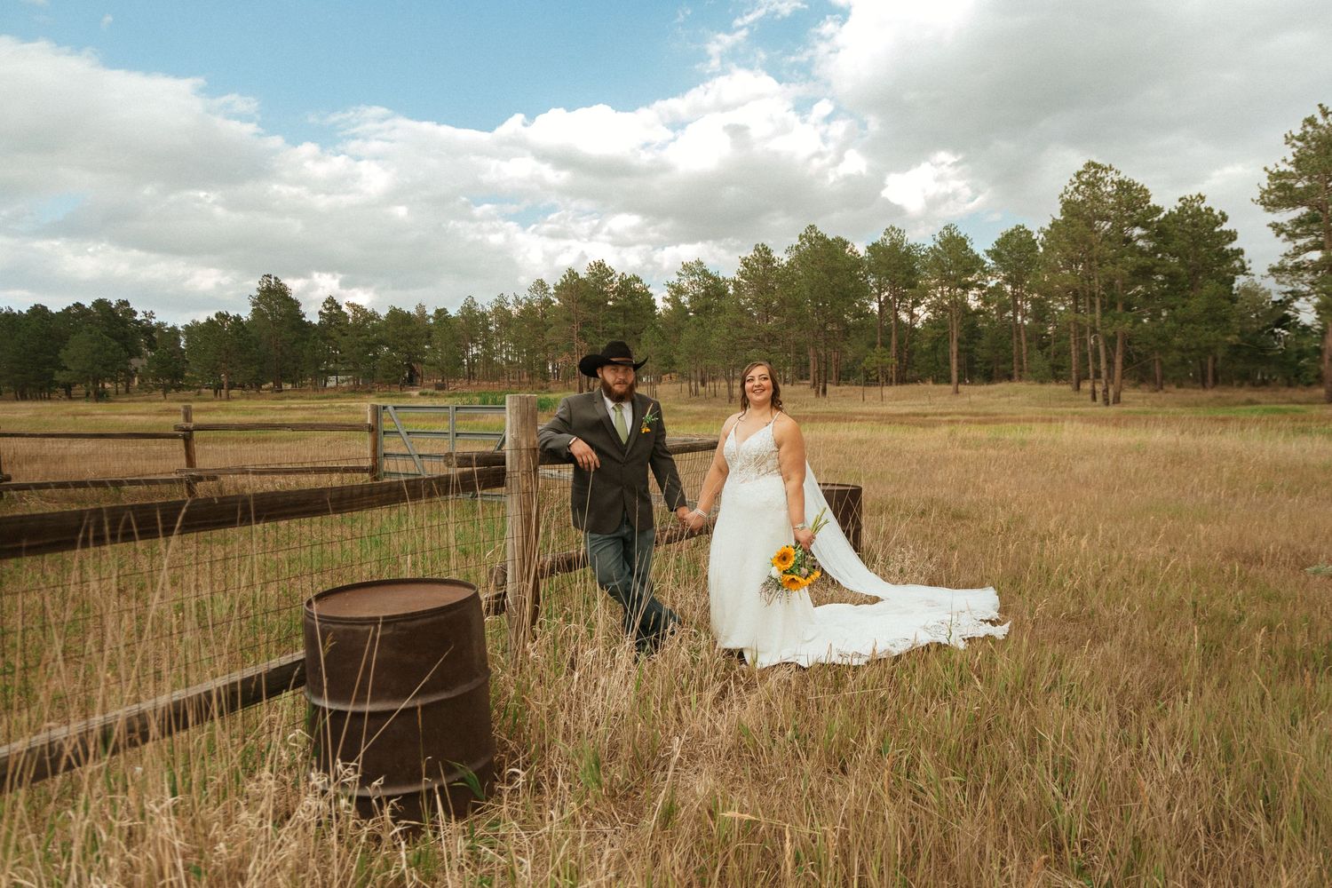 Two people in wedding attire stand in a rustic outdoor field setting with pine trees and wooden barrels at sunset.