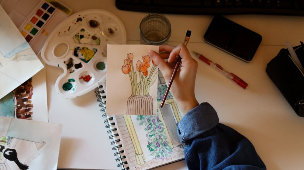 Overhead view of hands painting watercolor flowers in a sketchbook with art supplies.