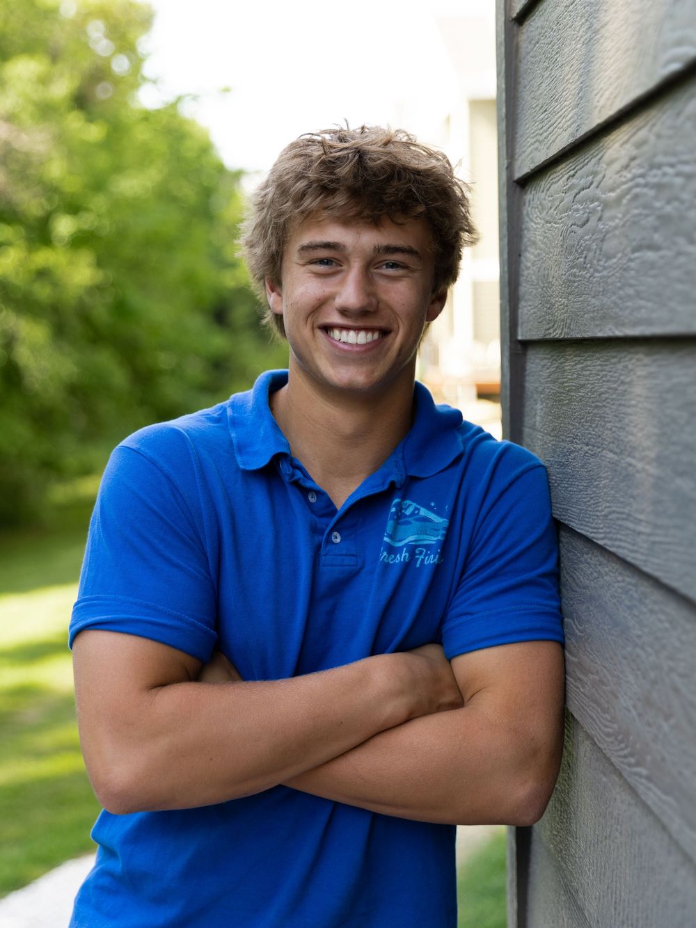 Smiling young man in blue polo shirt leaning against gray siding with arms crossed outdoors.