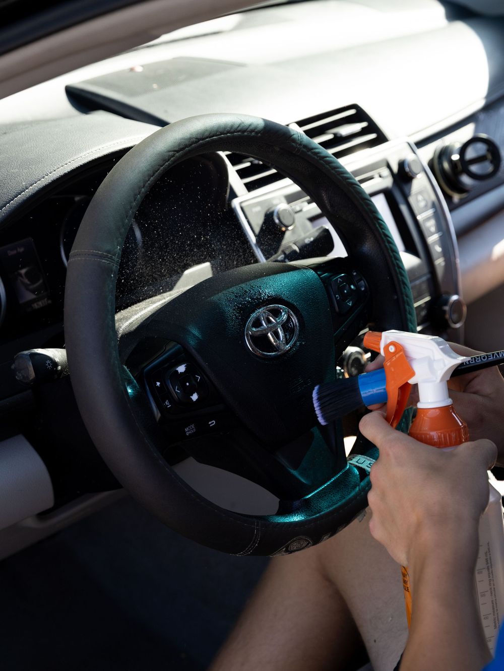 Person cleaning a Toyota car interior steering wheel with an orange spray bottle for detailing.