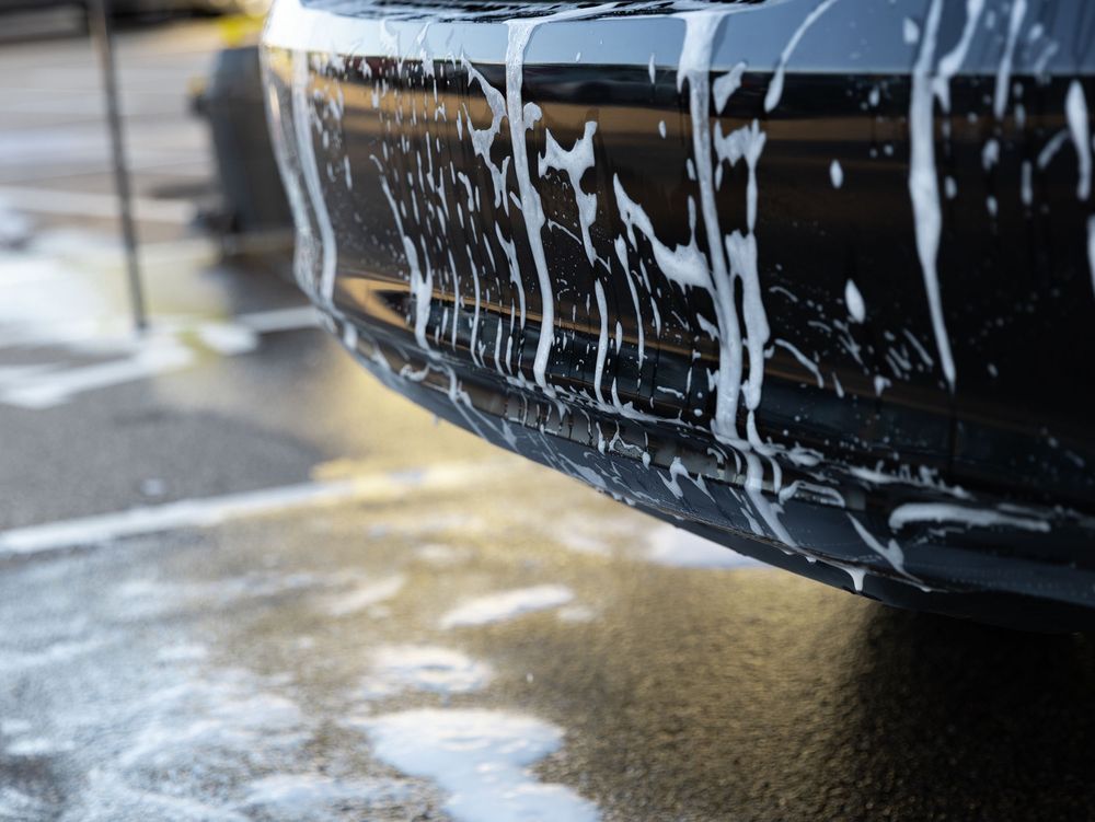 A dark car being washed with white foam soap suds dripping down the bumper on a wet surface.