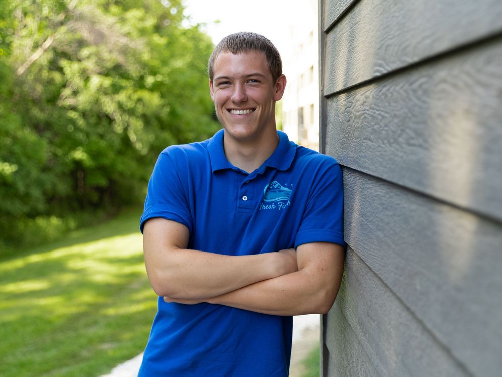Smiling young man in blue polo shirt leaning against gray siding with arms crossed outdoors.