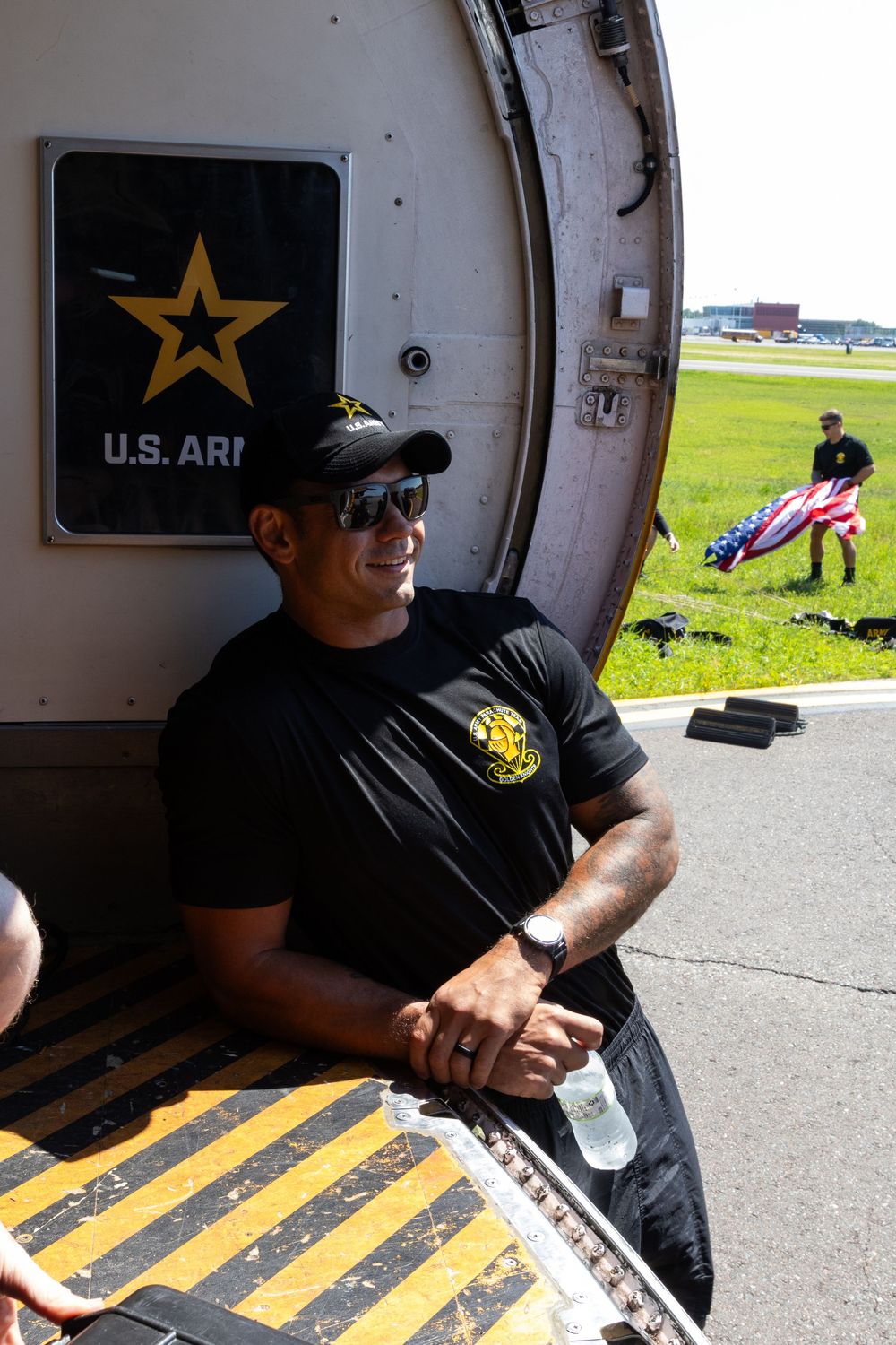A military personnel sits at the cargo door of an aircraft with a U.S. Army logo while someone carries gear on the airfield.