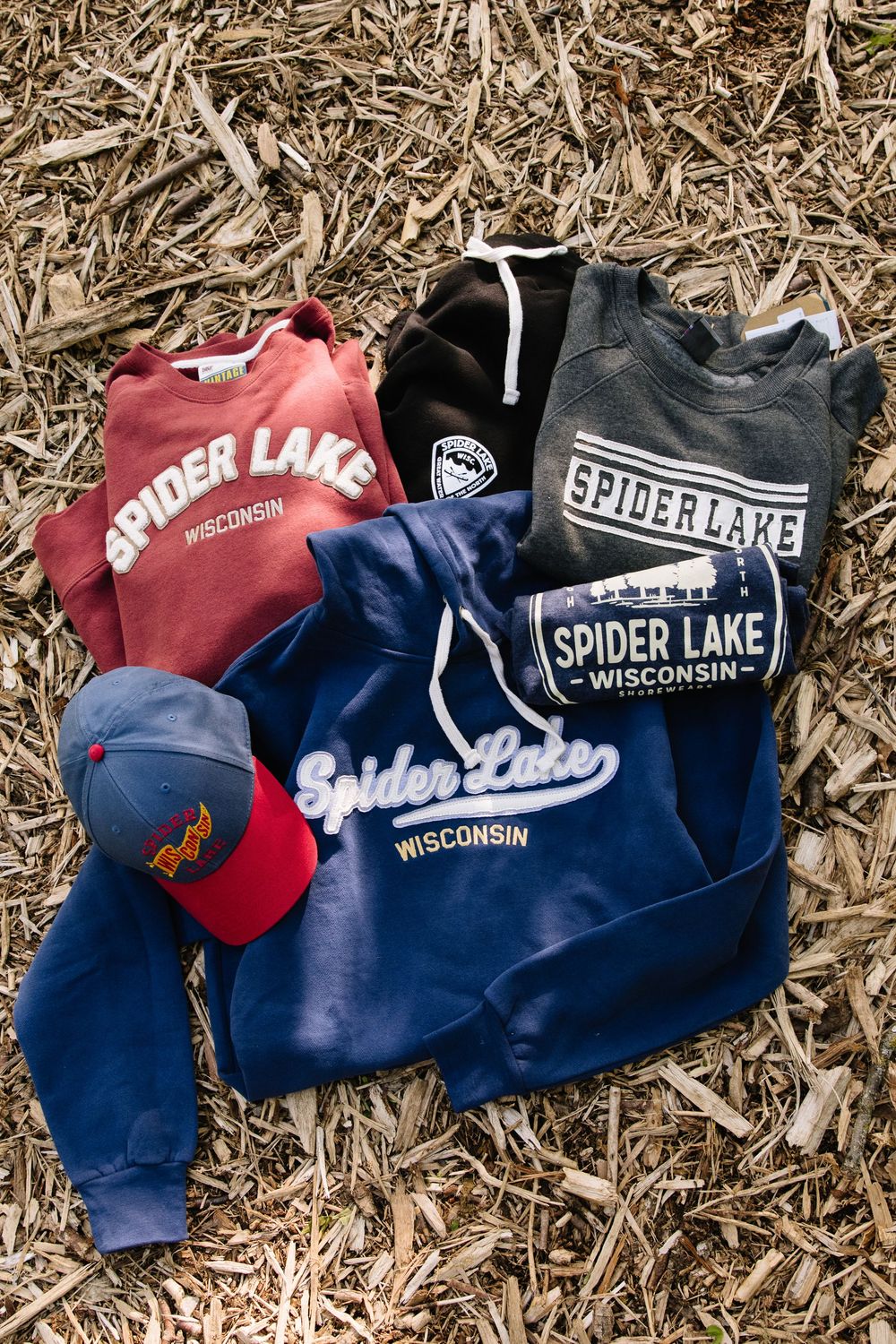 Collection of Spider Lake Wisconsin branded apparel including sweatshirts and baseball cap laid on dried grass background.