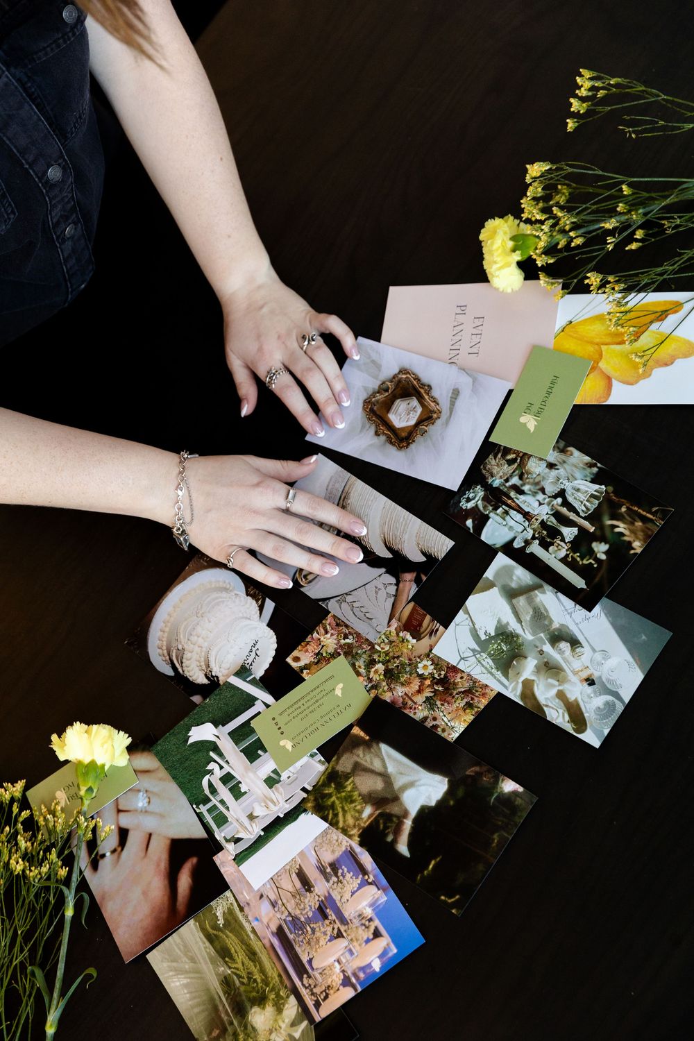 Hands arranging a collection of photographs and flowers on a dark surface in a memory keeping layout.