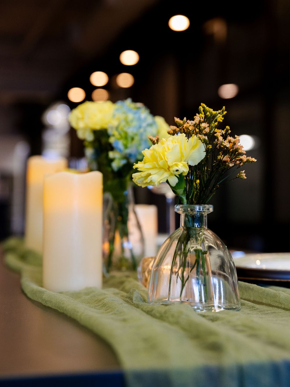 White candles and yellow flowers in glass vases line a sage green table runner creating an elegant centerpiece display.