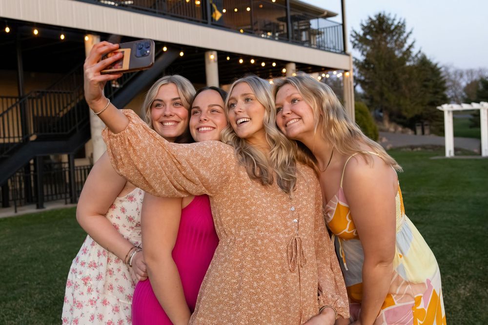 A group in colorful dresses smiles together while taking a selfie at an outdoor event.