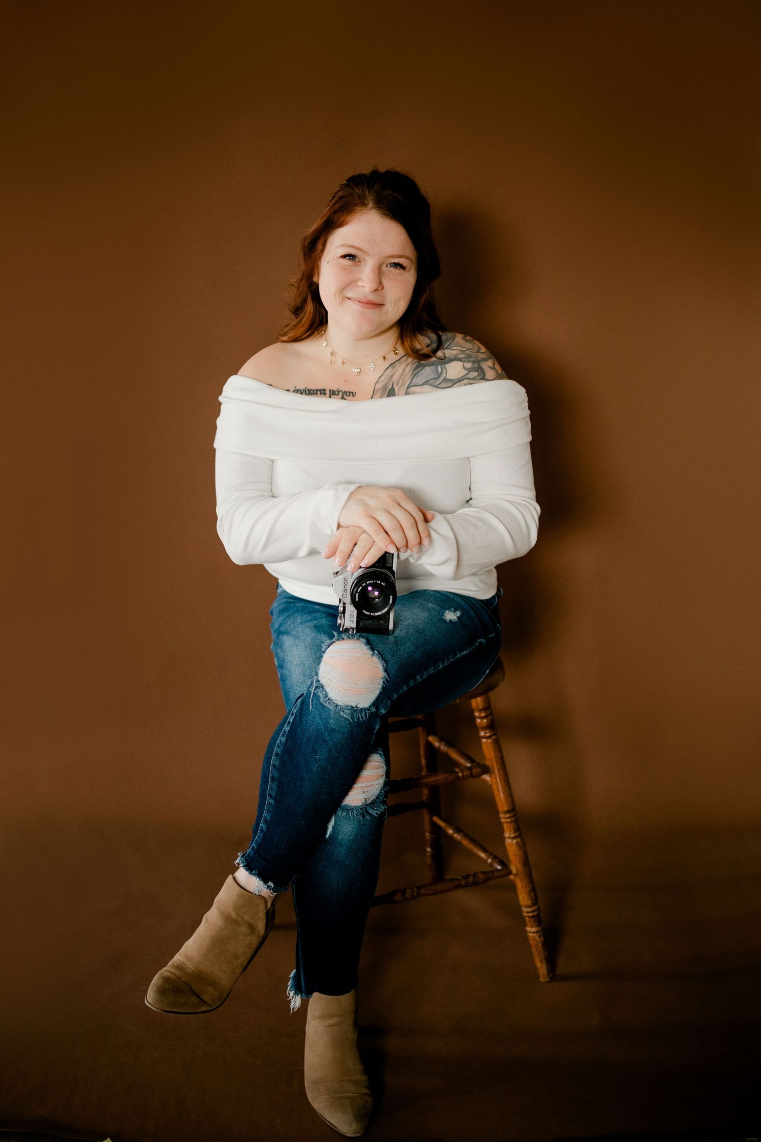 Smiling woman in white off-shoulder sweater and ripped jeans holding vintage camera on wooden stool.