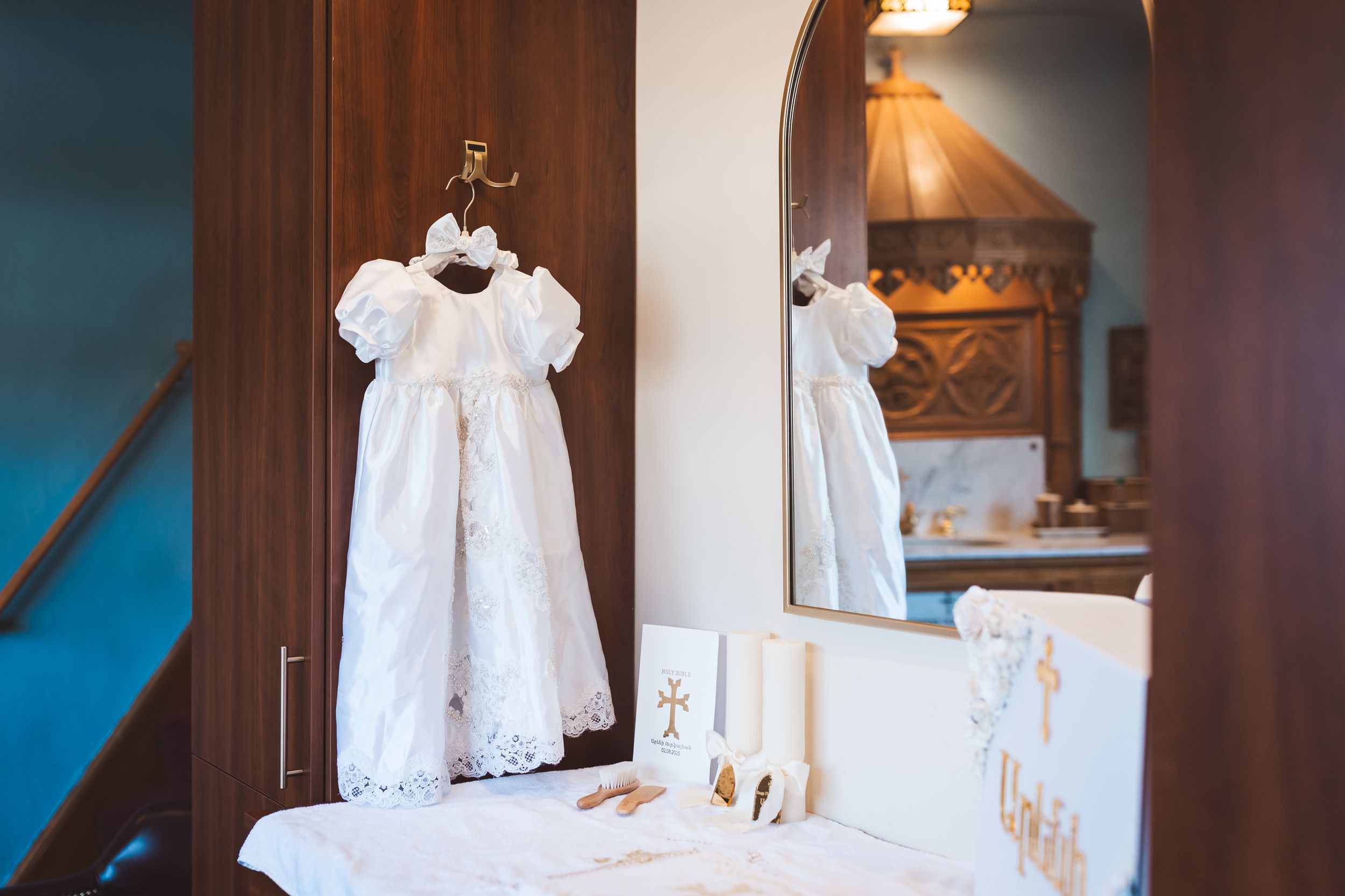 White christening gown hanging in room with mirror reflection and baptism accessories on table.