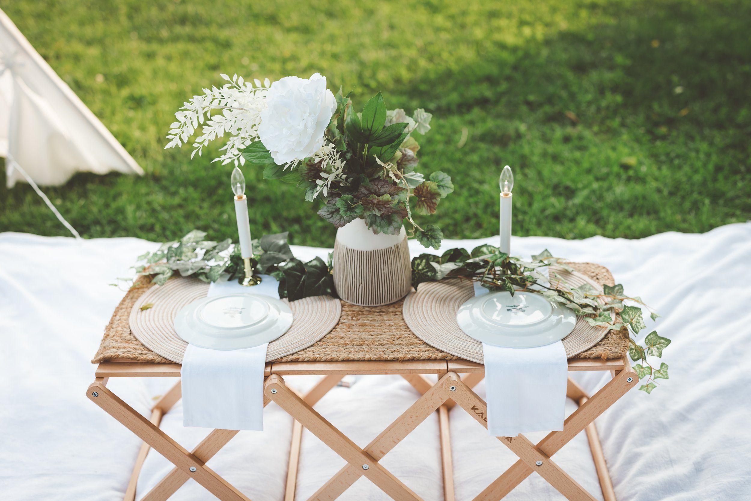 Elegant outdoor wedding table setting with white florals and greenery on a folding rattan table.