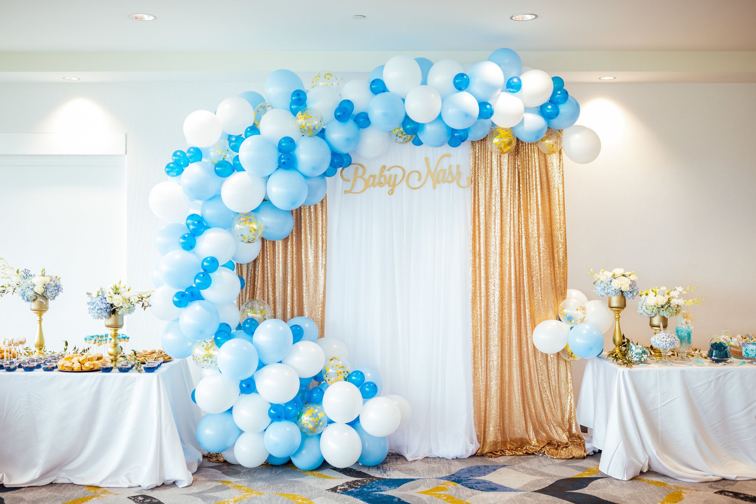 Blue white and gold balloon arch decoration over dessert table at elegant baby shower celebration.