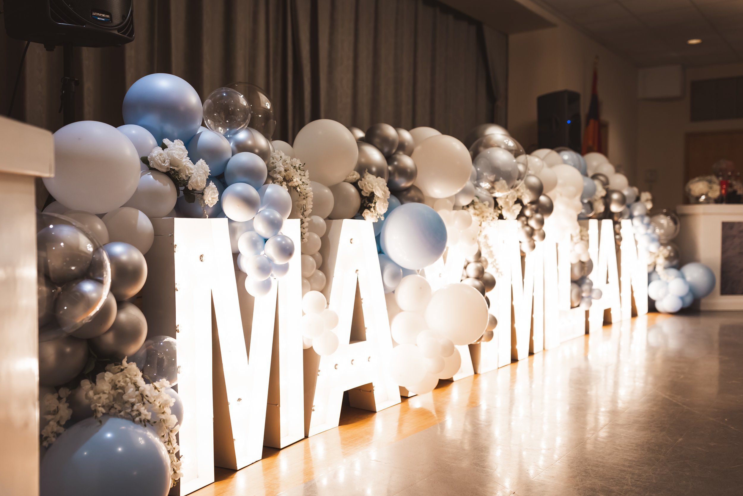 Large illuminated marquee letters spelling a name decorated with blue and silver balloons at a party.
