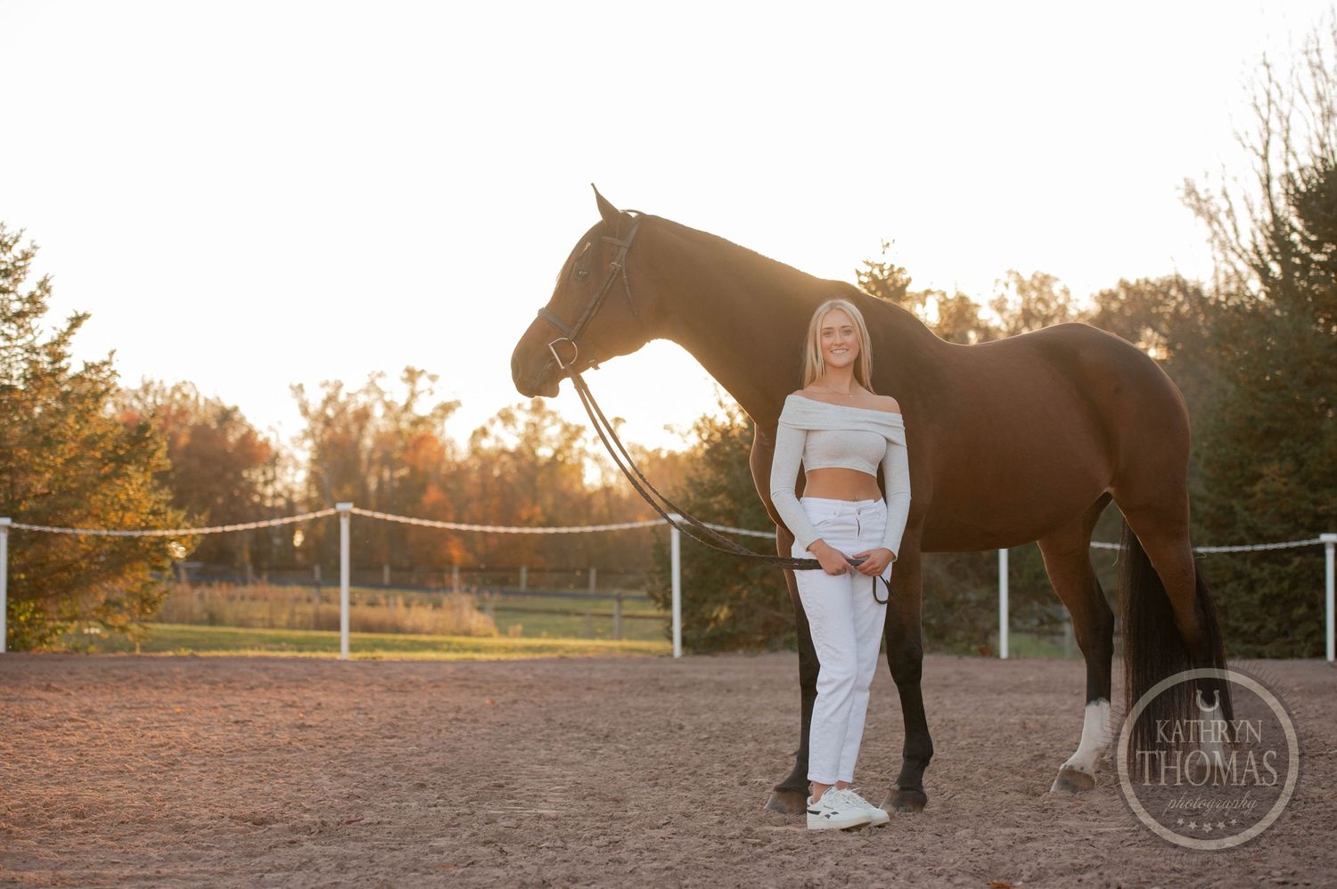 A bay horse stands quietly beside someone wearing white in the golden evening light.
