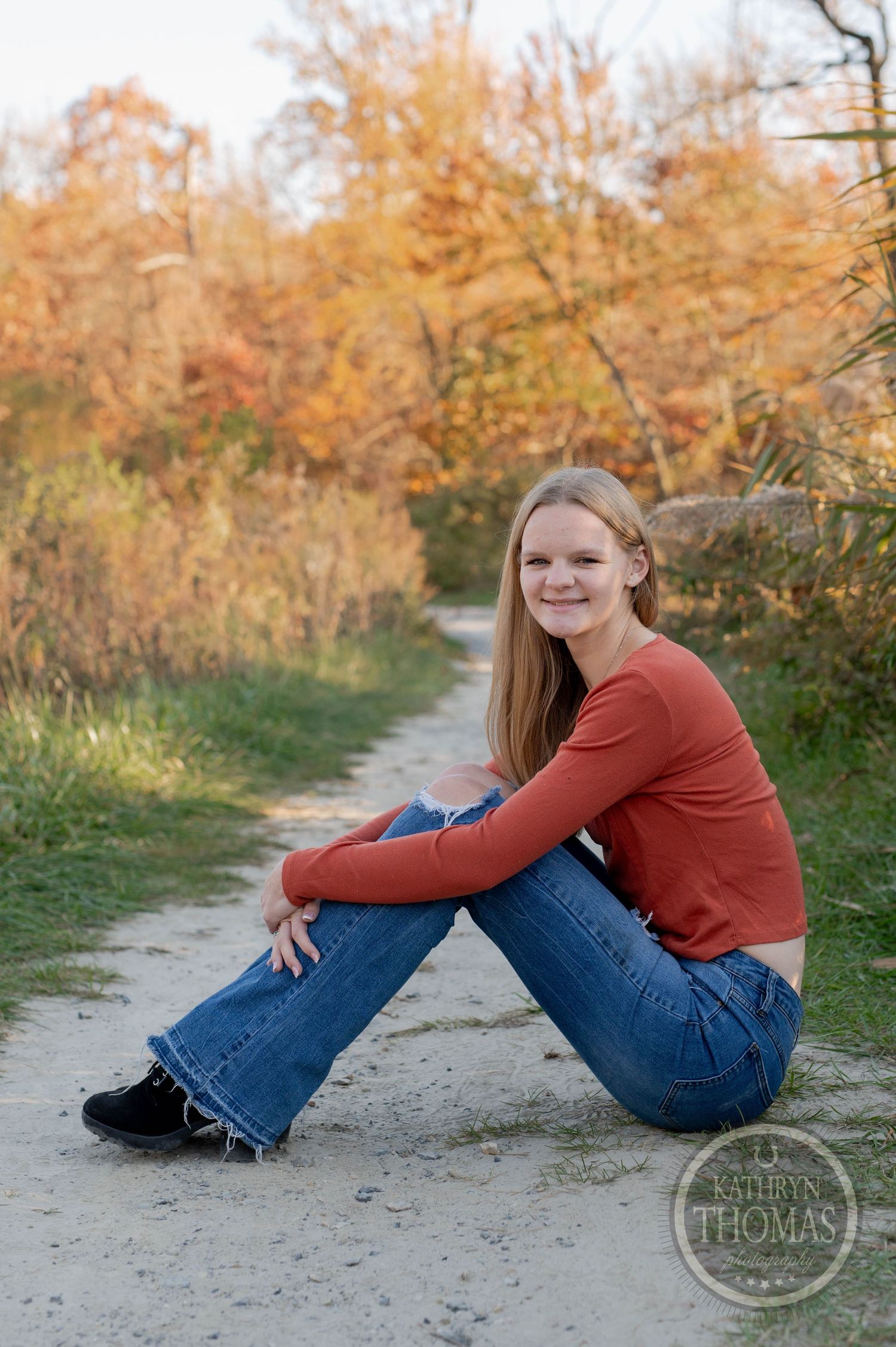 Person in rust-colored sweater sitting on path with autumn trees in background.