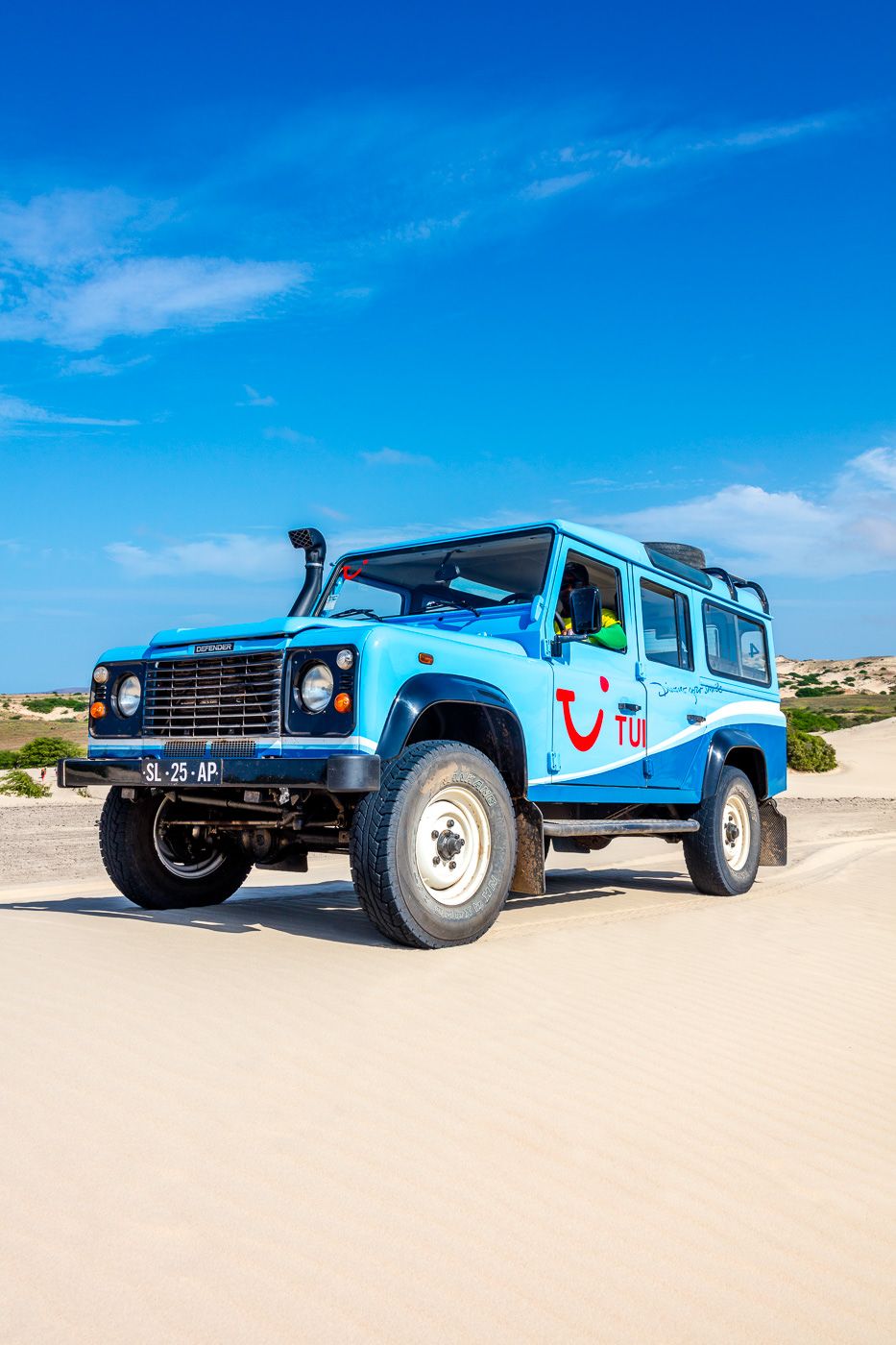 A bright blue Land Rover Defender parked on a sandy beach under clear blue skies with TUI tourism branding.