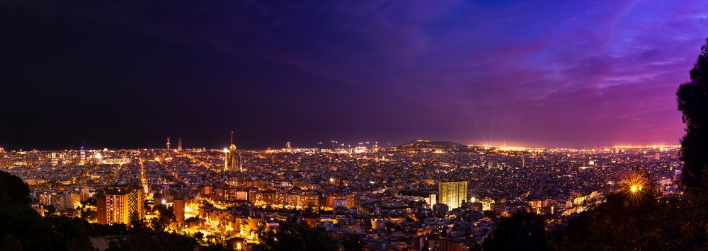 Panoramic cityscape of Barcelona at night with illuminated buildings and streets against a purple twilight sky.