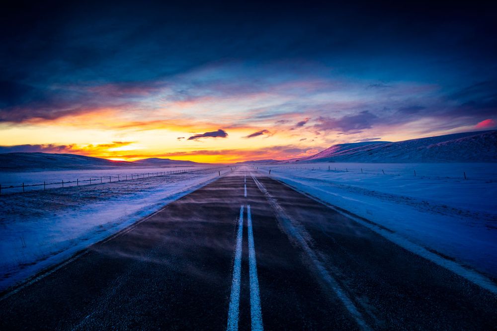 Empty winter highway stretches towards vibrant sunset with snow covered fields and dramatic colorful sky.