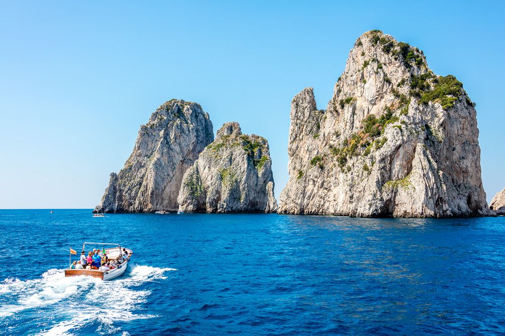 A small boat speeds past the famous Faraglioni rock formations rising from bright blue waters near Capri, Italy.