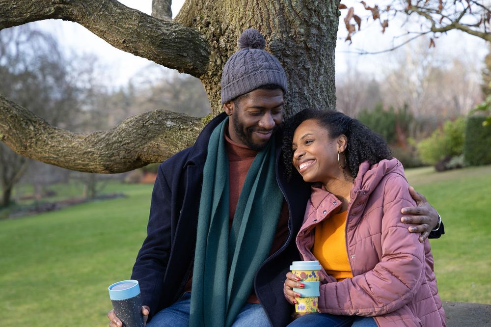 Two people sitting together outdoors under a tree branch sharing snacks and drinks on a sunny afternoon.