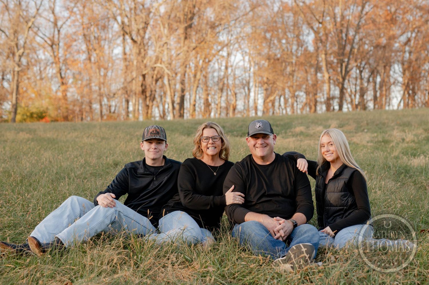 Family group lounging together on grassy hillside with autumn trees behind them.