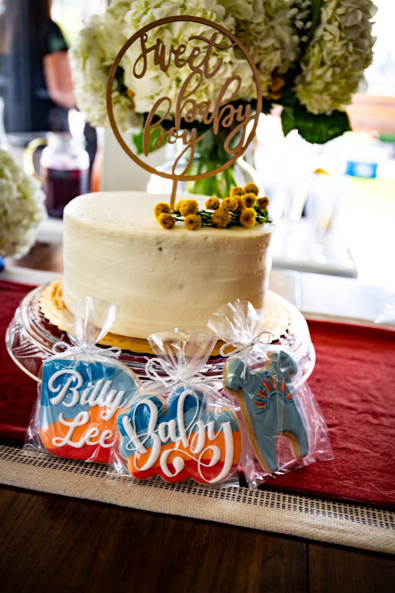 White baby shower cake with gold topper and decorated cookies wrapped in cellophane below.