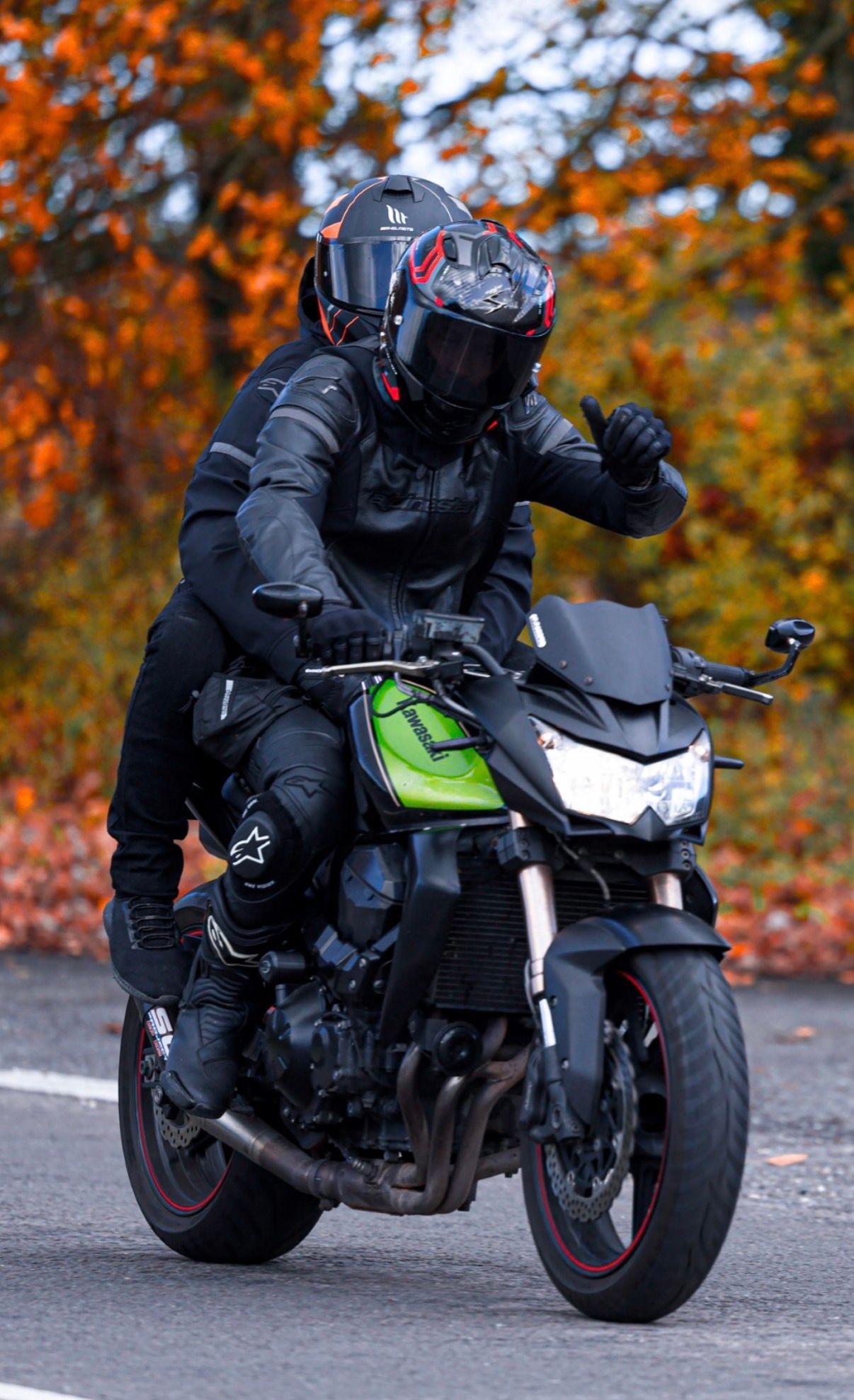 Motorcyclist in black gear rides a green and black sports bike against vibrant autumn foliage background.