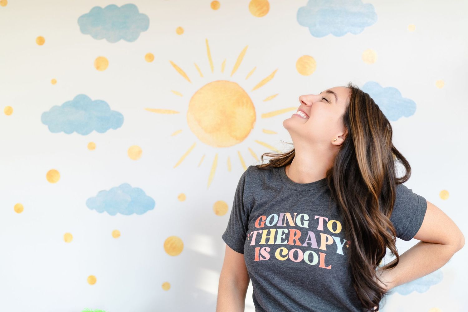 Woman in therapy school t-shirt laughing against wall with sun and cloud decorations.