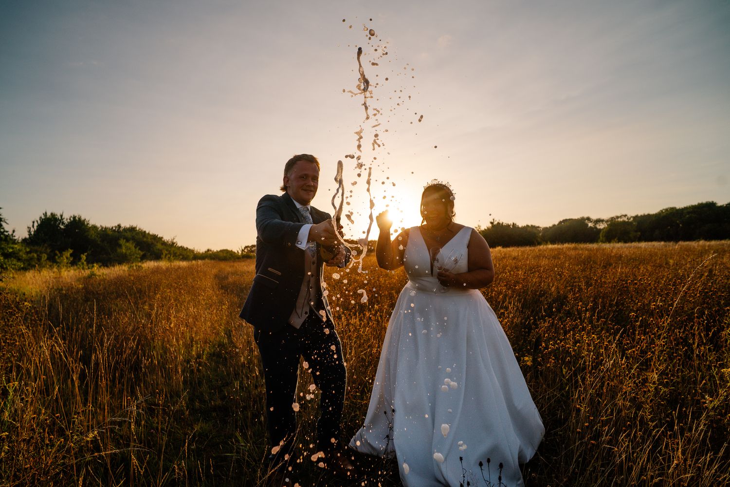 Couple popping champagne during their wedding photography and film investment celebration. Wide shot both very happy at sunset.