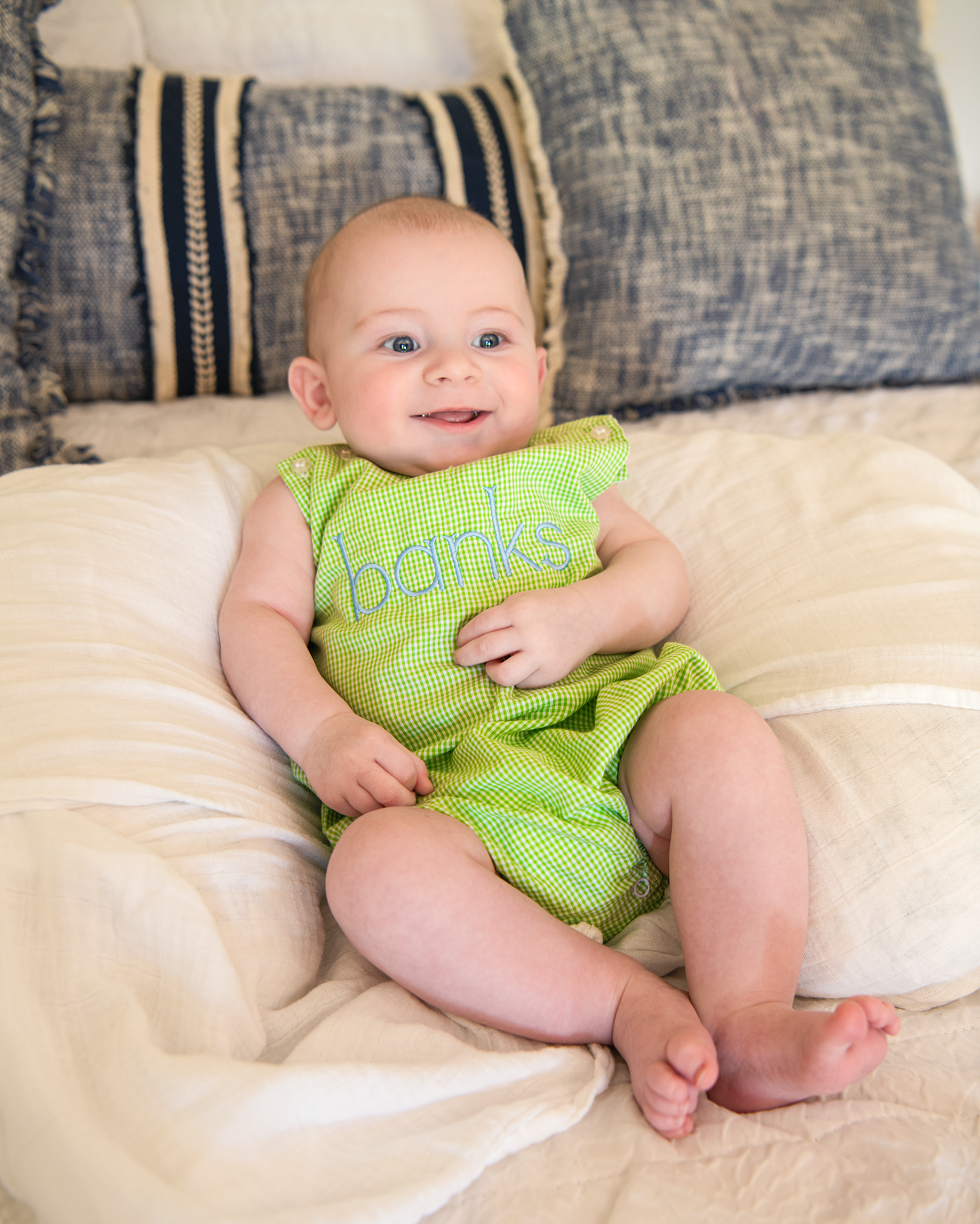 Smiling baby wearing a green romper with the name Banks embroidered on it, sitting on a bed.