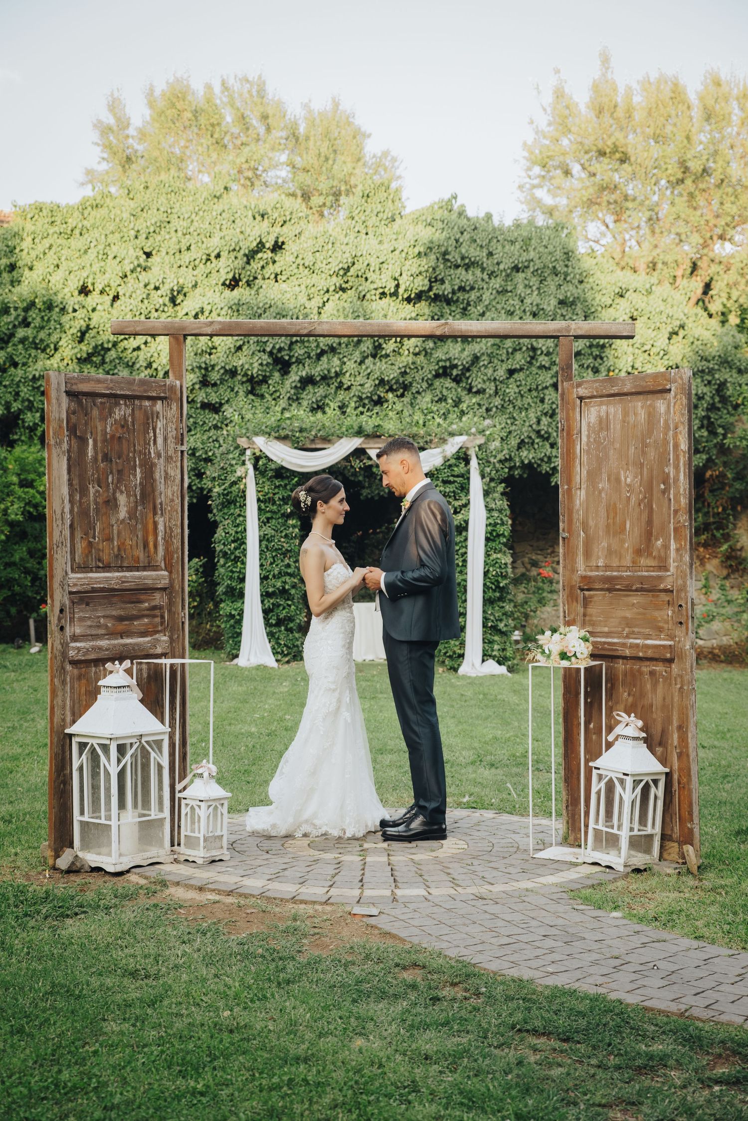A rustic outdoor wedding ceremony setup with vintage wooden doors, white lanterns, and a stone pathway in a garden setting.