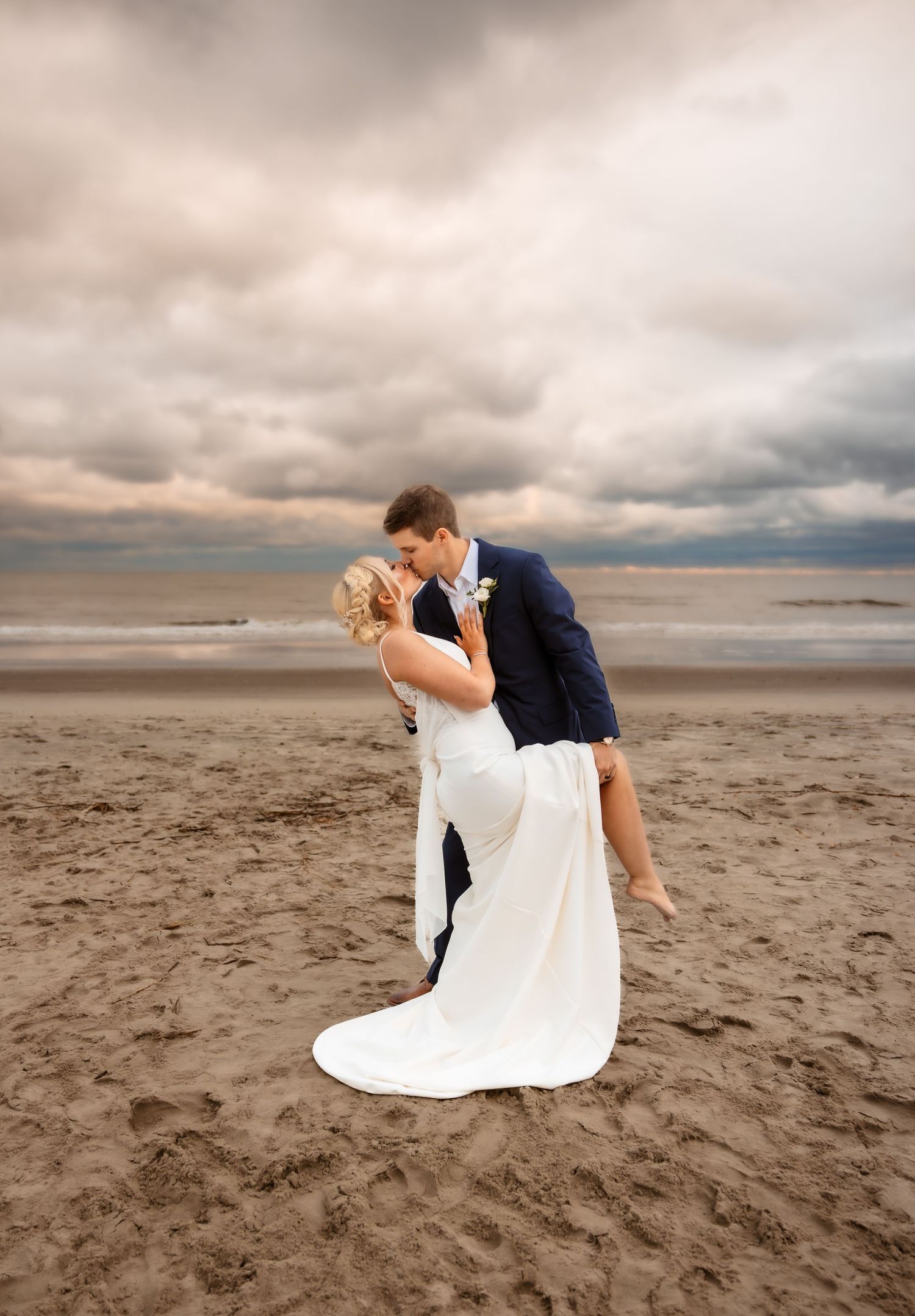 A romantic couple in wedding attire embrace and kiss on a moody beach with dramatic cloudy skies at sunset.