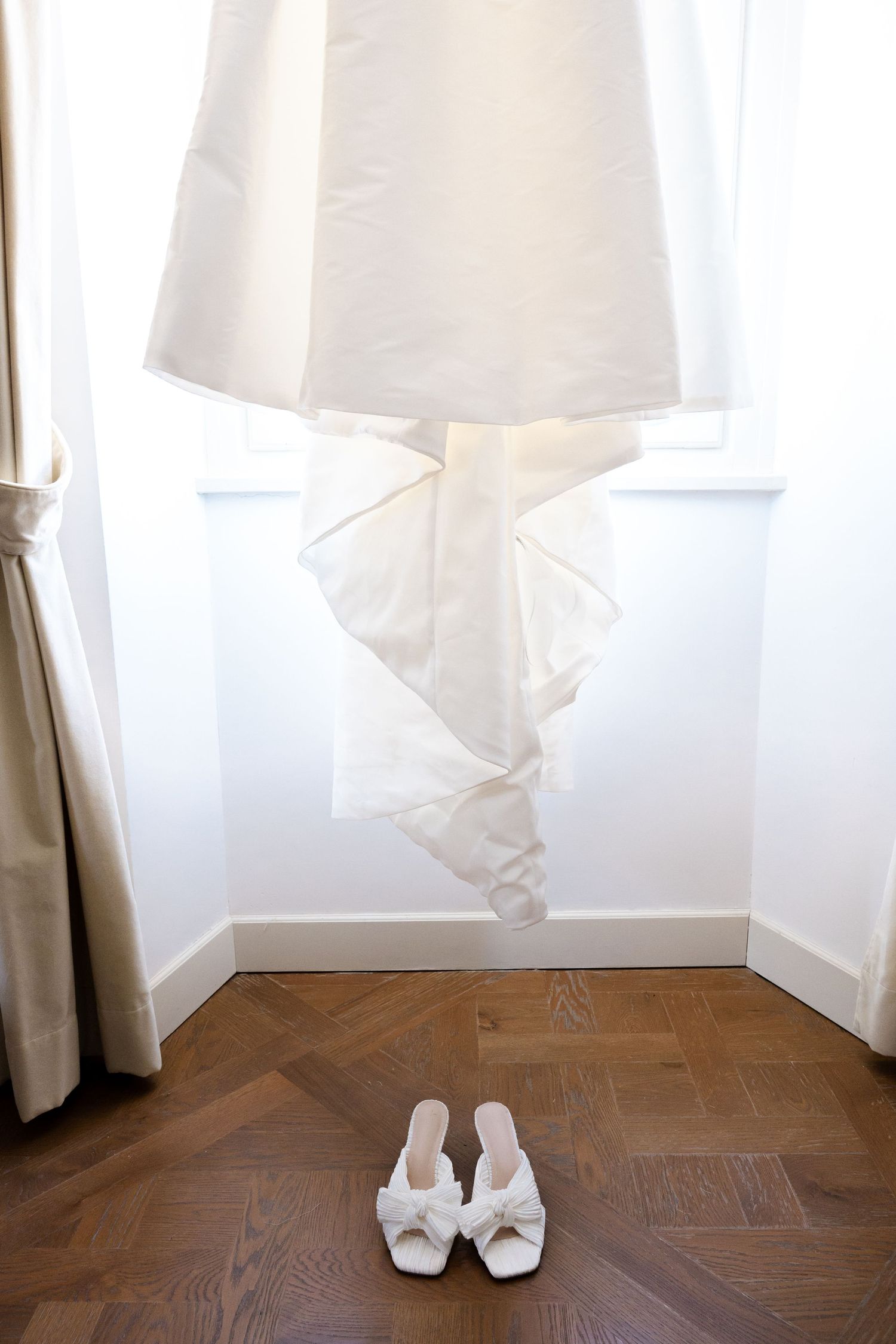 White curtains billow softly in a room with herringbone wood flooring and a pair of white slippers by the window.