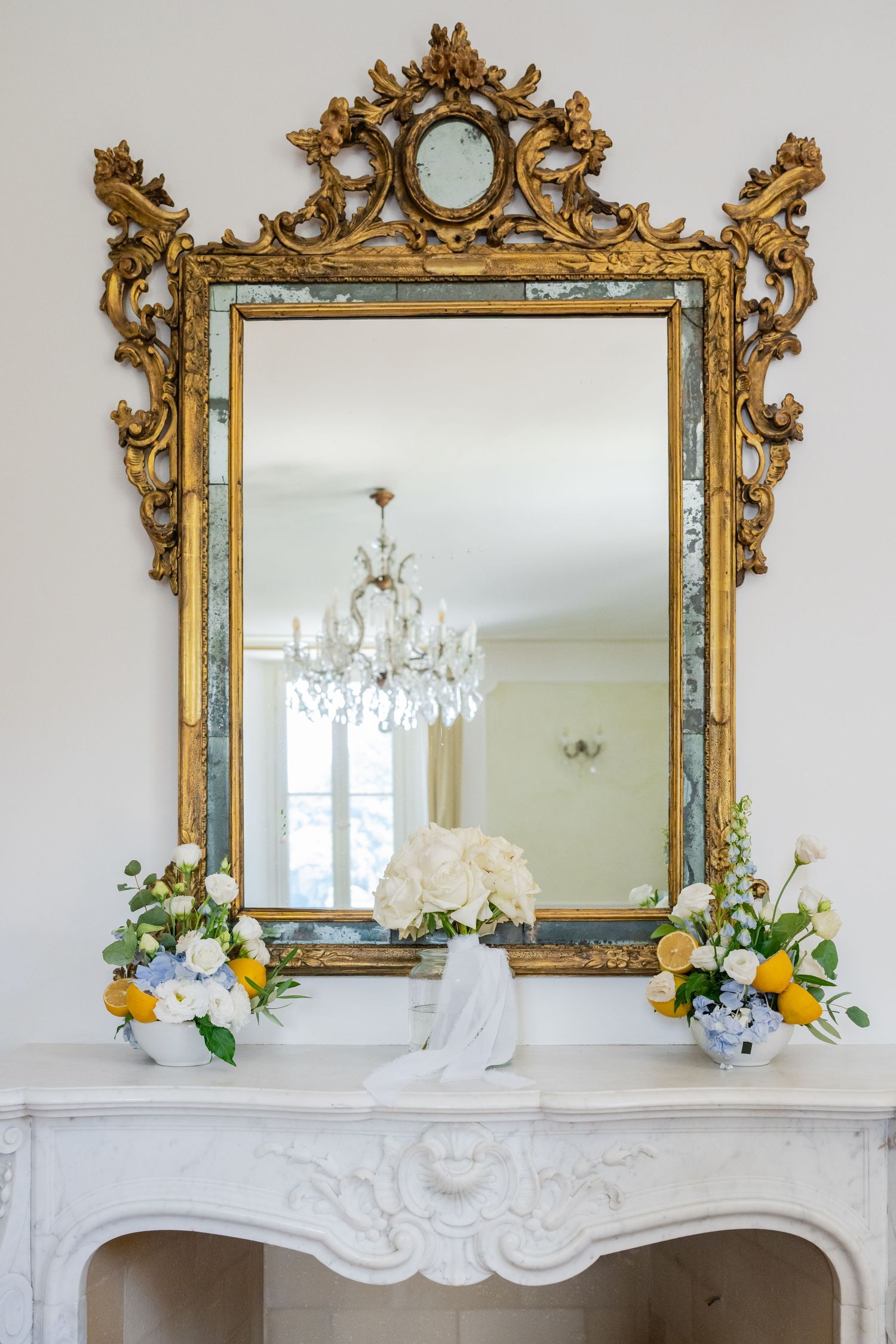 Ornate gold baroque mirror above white marble fireplace mantel with crystal chandelier reflection and floral decorations.
