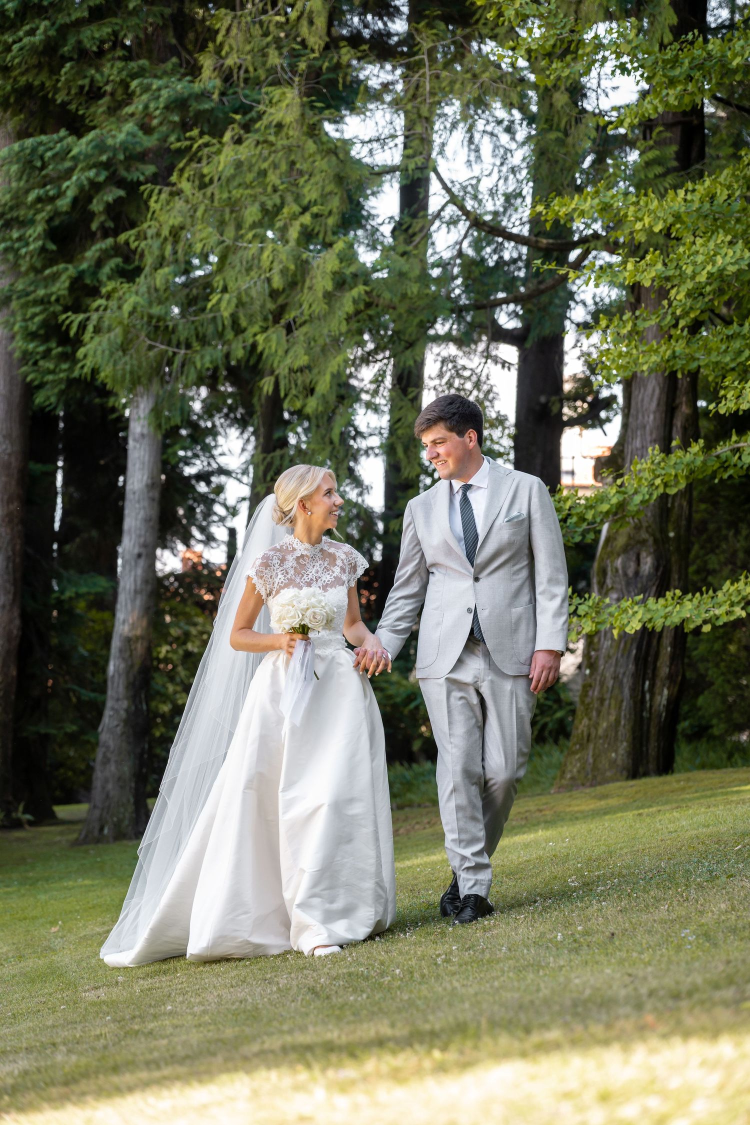 Wedding party in light gray suits and white dresses walking down a grassy hill among tall evergreen trees.