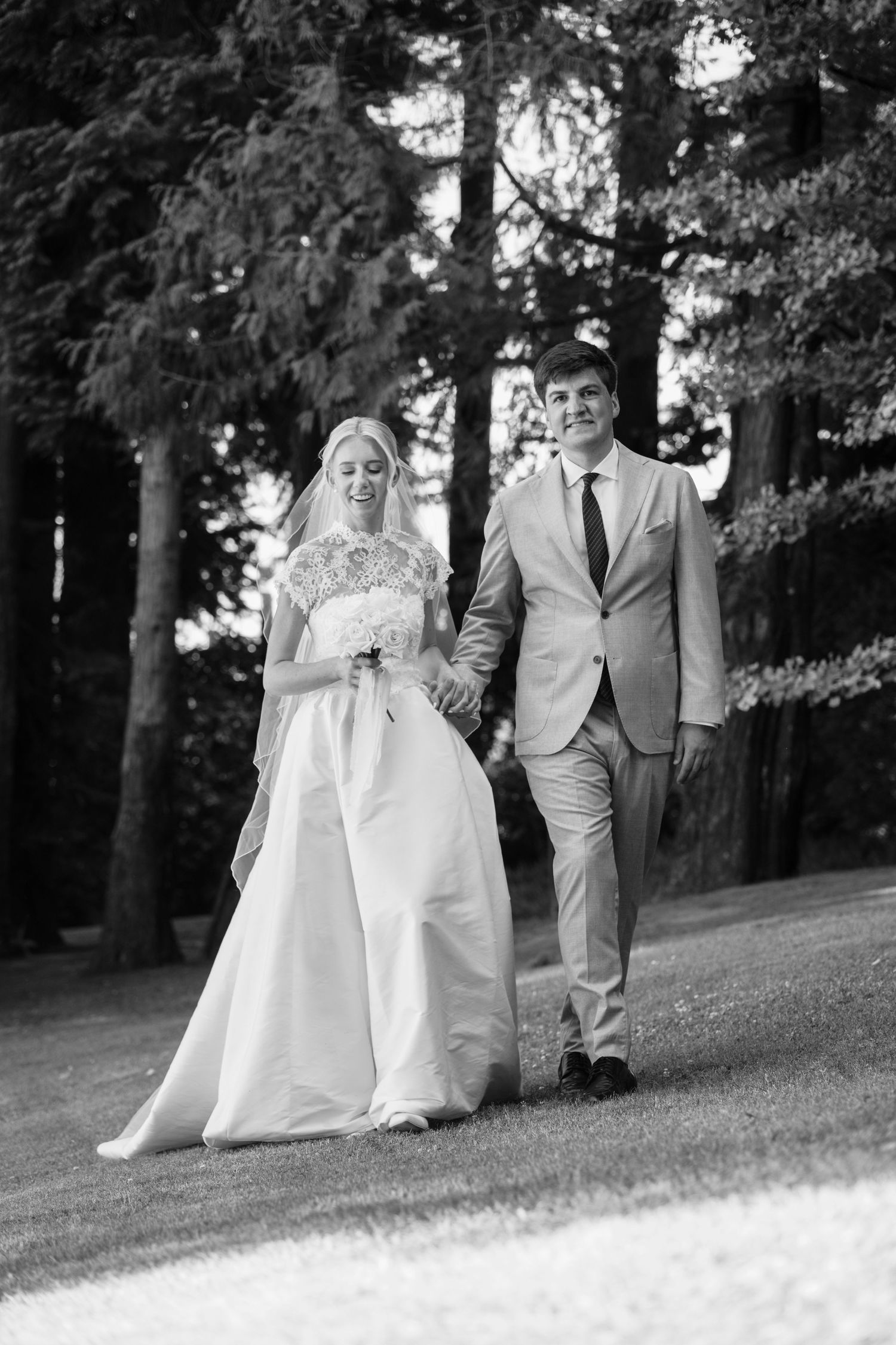 A black and white wedding photo showing a couple walking together outdoors among tall trees.