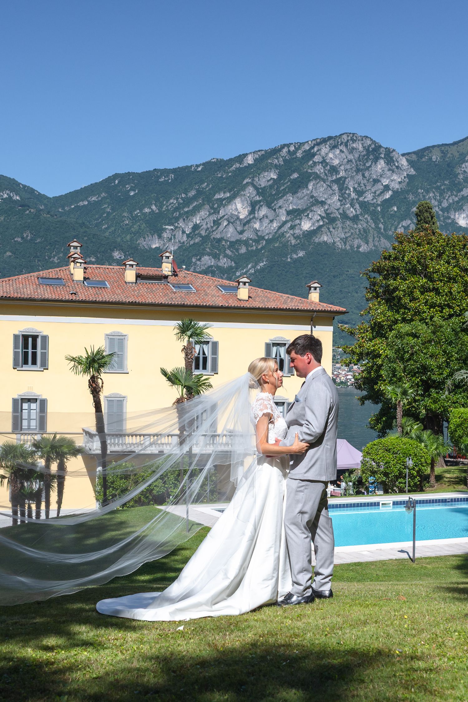 A wedding couple poses for romantic photos on a lawn with mountains and a villa with pool in the background.