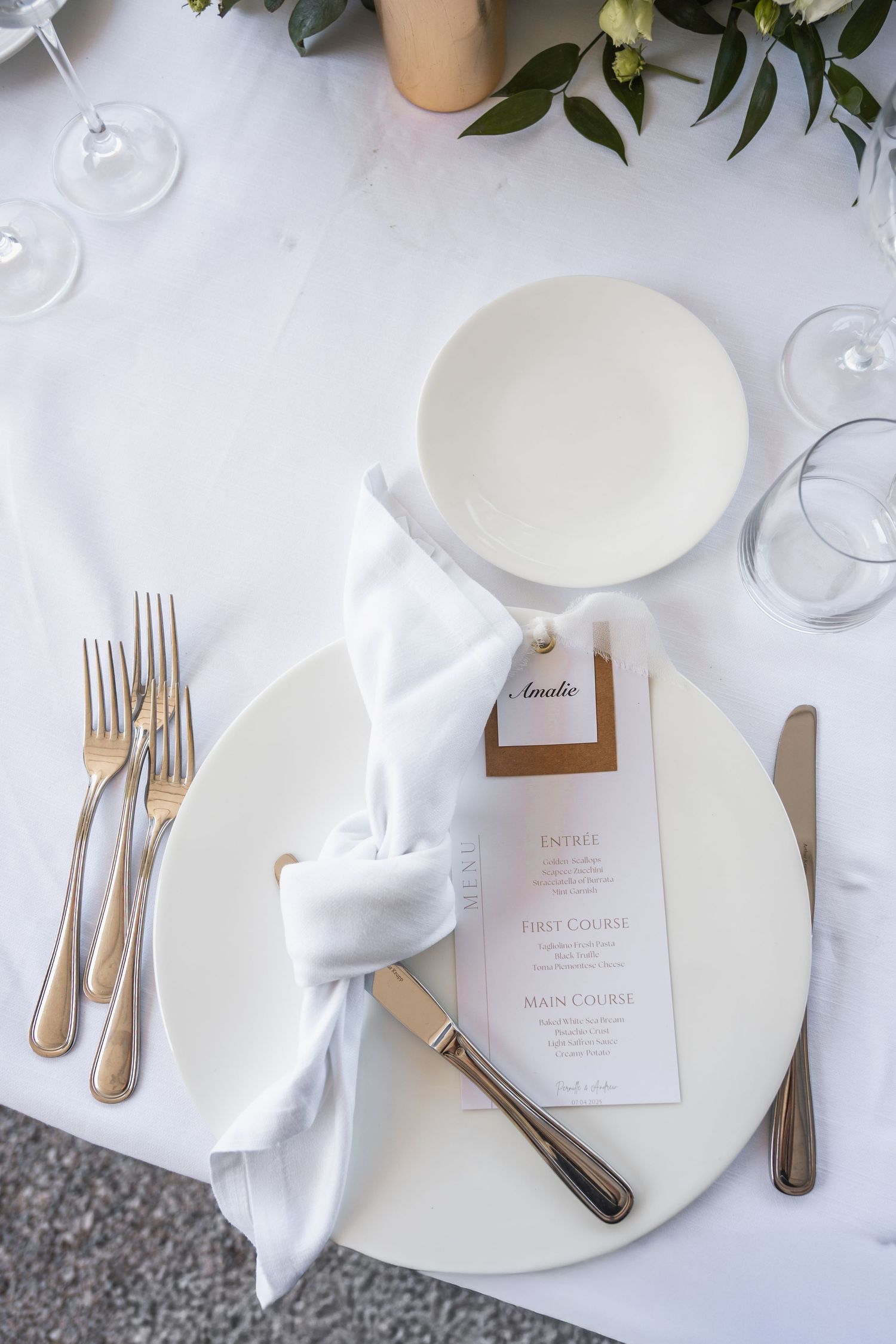 Elegant place setting with white plate, gold cutlery, folded napkin and menu card on crisp white tablecloth.