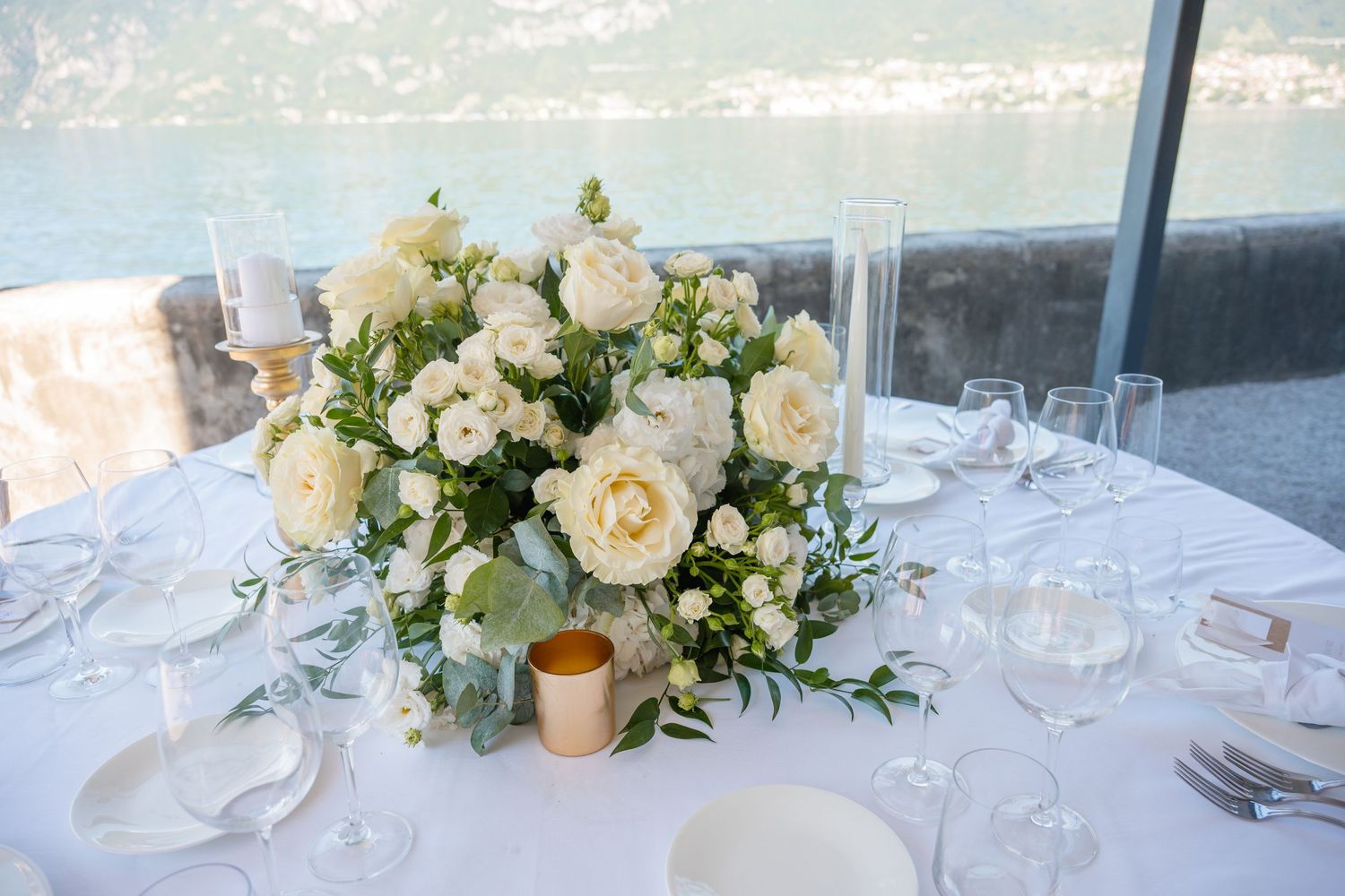 Elegant wedding table setting with white rose centerpiece and gold candle holder on white linen tablecloth.