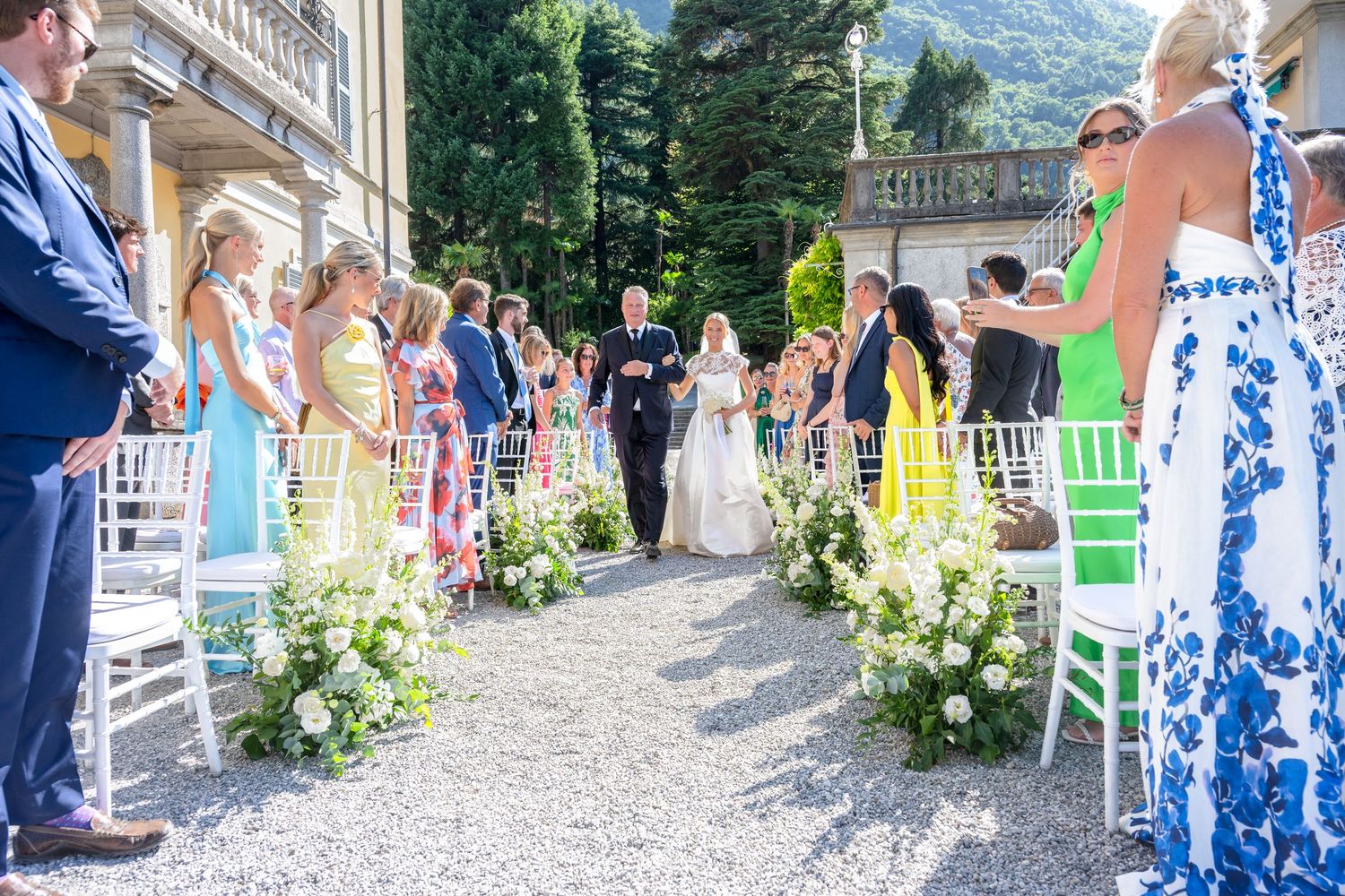 Wedding guests seated in white chairs along a garden pathway with floral arrangements and mountains in the background.
