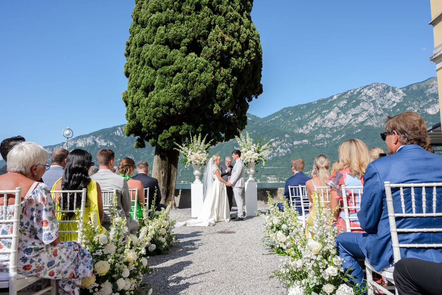 Wedding guests seated in white chairs at an outdoor ceremony overlooking Lake Como and mountains in Italy.