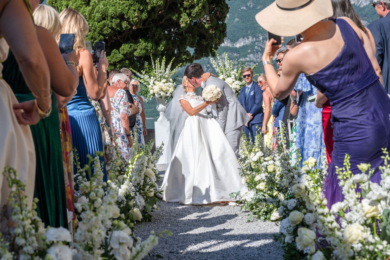 A group of wedding guests gathered outdoors in formal attire surrounded by floral decorations and greenery.