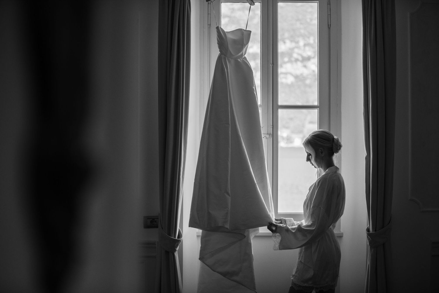 A black and white photo shows a silhouette of someone looking through a window with curtains.