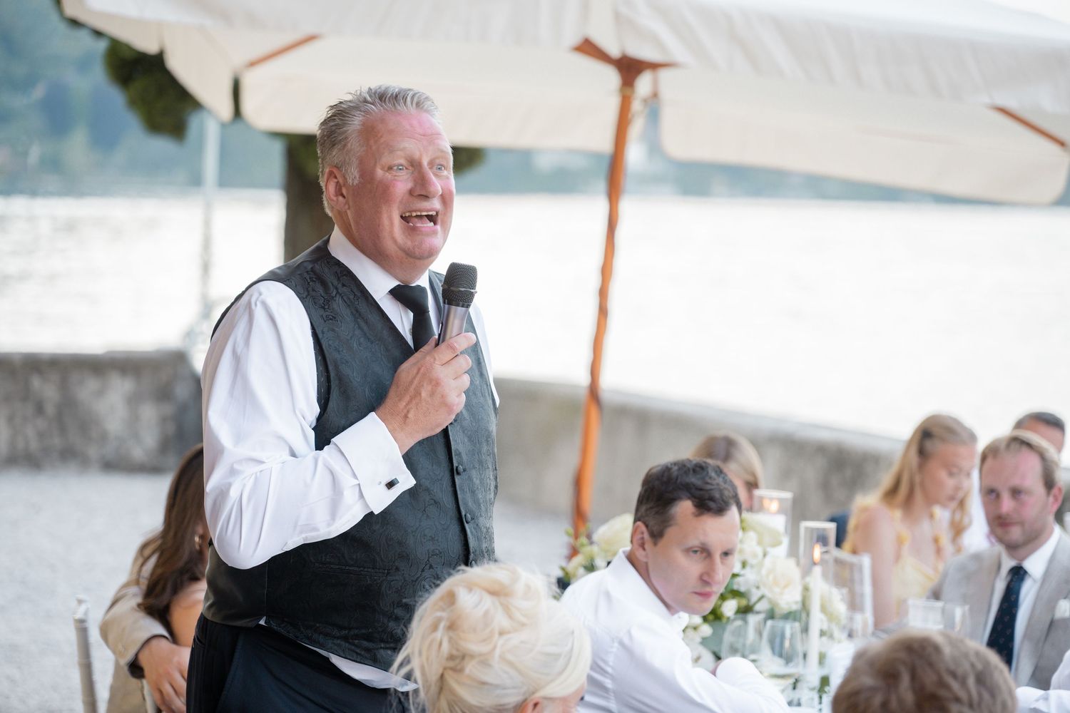 Wedding guests seated at tables under a white tent laugh and socialize during an outdoor reception celebration.